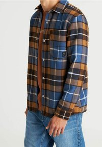 Koszula flanelowa w niebiesko-brązowo-czarnej kratę. Posiada zapinany na guziki przód, jedną kieszeń na piersi oraz długie rękawy. Nosi się ją na brązową koszulę.