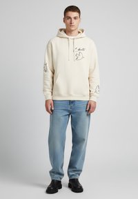 Felpa beige con tasca frontale, cappuccio con coulisse e disegni di uccelli neri stampati sul petto e sulle maniche, abbinata a jeans azzurri chiari e stivali neri.
