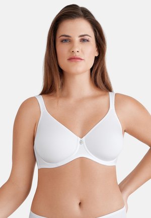 Vrouw met lang bruin haar, gekleed in een witte beugelbeha, staand met één hand op haar heup tegen een effen achtergrond.