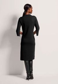 Schwarzes Strickkleid mit dreiviertelärmeln, knielanger Schnitt und taillierter Silhouette. Kombiniert mit kniehohen schwarzen Stiefeln.