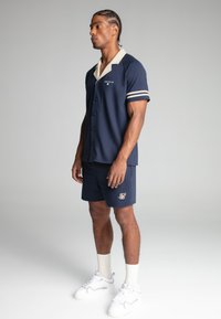 Marineblauwe korte mouwen shirt met een crèmekleurige kraag en gestreepte mouwen, gecombineerd met marineblauwe shorts met een gouden logo, gedragen met witte sneakers.