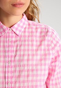 Růžová a bílá kostkovaná košile na knoflíky s klasickým límcem, která má texturovanou tkaninu a jemné pomačkání. Viditelné dekorační knoflíky.