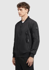 Schwarze Bomberjacke mit Reißverschluss vorne, gerippten Bündchen, zwei seitlichen Reißverschlusstaschen und einer aufgenähten Tasche am Ärmel. Glattes, leichtes Material.