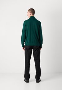 Maglione verde a maniche lunghe con colletto alto, aderente in vita, abbinato a pantaloni neri e scarpe nere, visto da dietro.