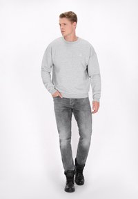 Grijze sweatshirt met ronde hals en lange mouwen, gecombineerd met vervaagde grijze jeans en zwarte enkelboots. Subtiel ankerdetail op de trui.