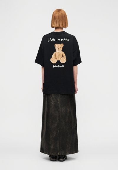 Μαύρο φαρδύ μπλουζάκι με γραφικό αρκουδάκι σε ταμπά χρώμα στην πλάτη, με τα κείμενα «BEAR IN MIND» και «Palm Angels» σε λευκό.