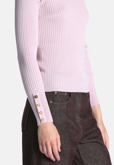 Femme portant un pull côtelé rose pâle avec des boutons dorés sur le poignet de la manche et un pantalon taille haute marron foncé.