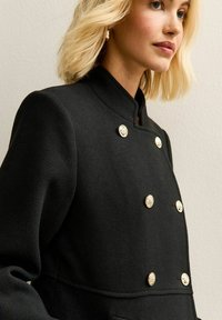 Zwarte dubbelknopsgesp coat met hoge kraag, textuurstof en gouden decoratieve knopen. Inclusief voorzakken voor extra functionaliteit.