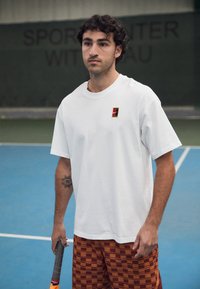 Maglietta atletica bianca con un piccolo logo multicolore sul petto, abbinata a pantaloni corti a quadri marroni. La persona tiene una racchetta da tennis.