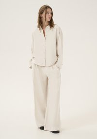 Blusa di seta beige con bottoni, vestibilità comoda. Abbinata a pantaloni beige a gamba larga, entrambi presentano una consistenza liscia e morbida. Scarpe nere completano il look.