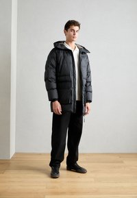 Schwarze gepolsterte Jacke mit Kapuze, ausgestattet mit einem Reißverschluss und seitlichen Taschen. Getragen über einem weißen Hemd, kombiniert mit schwarzen Hosen und Schuhen.