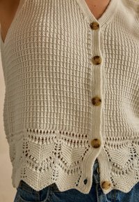 Strickweste in Off-White mit strukturiertem Muster. Verfügt über einen wellenförmigen Saum und Holzknöpfe, die gut zu eng anliegenden Jeans passen.