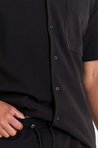 Zwart kantoorshirt met korte mouwen en een gestructureerde stof, voorzien van een borstzak en zwarte knopen, gedragen over zwarte joggers met een trekkoord.