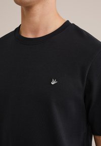 Vyras dėvi juodą marškinėlį su apvalia apykakle, ant kairiojo krūtinės pusės yra mažas balto paukščio logotipas.