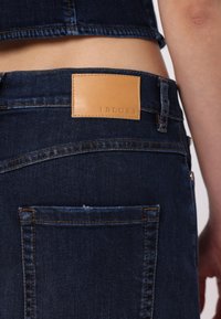 Mörkblå denimjeans med en tan läderlapp med "i BLUES" präglat, synlig sömnad och en bakficka på högra sidan.