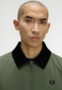 Giacca leggera verde oliva con colletto in velluto a coste nero, chiusura a zip e logo ricamato sul petto. Texture liscia.