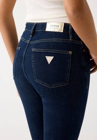 Jean en denim bleu foncé avec une coupe ajustée, des coutures dorées et une patch en forme de triangle avec le logo "GUESS" sur la poche arrière.