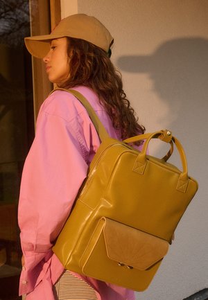 Femme portant une casquette beige et une chemise rose, porte un grand sac à dos jaune moutarde avec une poche avant, debout à l'intérieur près d'une porte en bois.