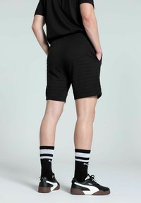 Shorts negri cu dungi orizontale, cu buzunare laterale, purtați cu adidași PUMA negri și albi și șosete cu dungi până la glezne.