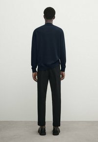 Marinefarbener Strickpullover mit rundem Ausschnitt, kombiniert mit schwarzen maßgeschneiderten Hosen. Glatte Textur mit lockerer Passform und umgeschlagenen Säumen. Schwarze Schuhe.
