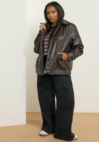 Giacca in similpelle marrone con zip frontale, ampio colletto e tasche laterali, abbinata a pantaloni neri a gamba larga e sneakers bianche.