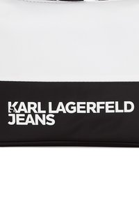 Juodas ir baltas nailono krepšys su ryškiu spausdintu tekstu "KARL LAGERFELD JEANS". Lygus paviršius, plokščia, stačiakampio formos ir užtrauktuku uždaromas.