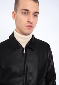 Veste en cuir noir avec fermeture éclair, dotée d'un col et d'une finition texturée. Le mannequin porte un pull à col roulé crème à côtes en dessous.