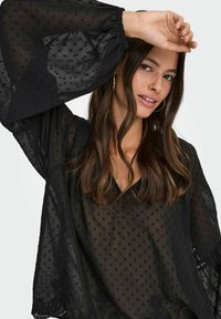 Blusa nera trasparente con pois texturizzati, scollatura a V e maniche a campana. Presenta ricami floreali sull'orlo. Tessuto leggero.