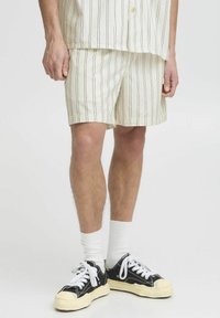 Kremfargede shorts med vertikale striper i mørkebrunt, laget av lett stoff. Kombinert med svarte sneakers og hvite ankelsokker.