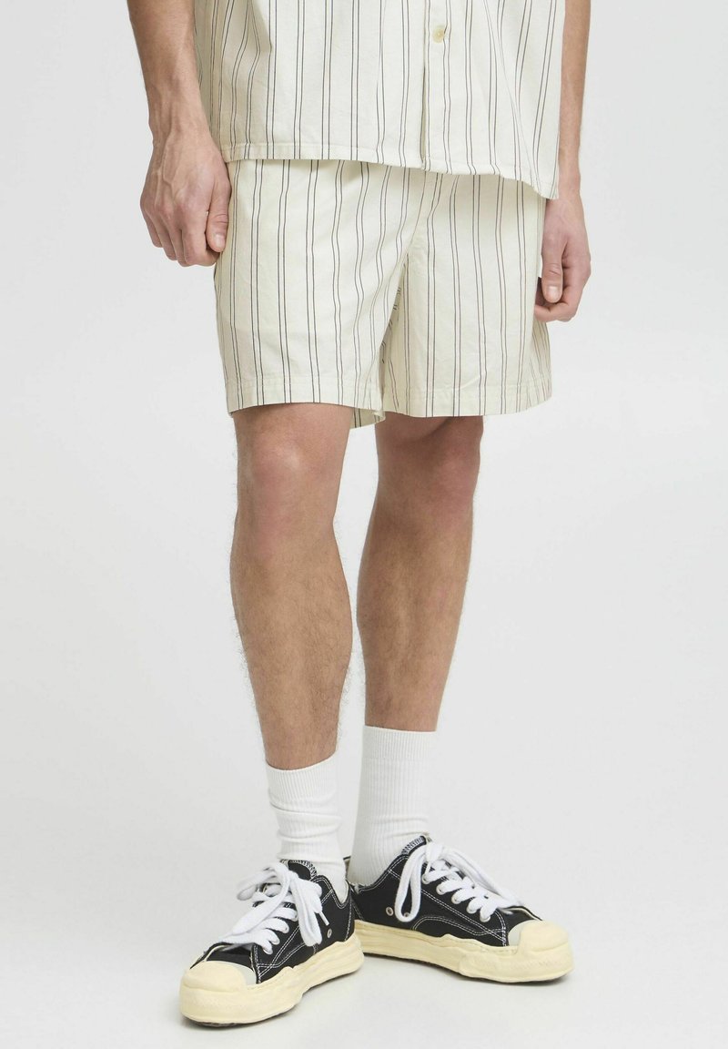 Cremefarbene Shorts mit vertikalen Streifen in Dunkelbraun, aus leichtem Stoff. Kombiniert mit schwarzen Sneakers und weißen Socken.