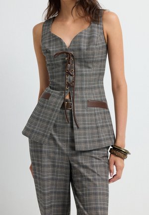 Femme portant un haut sans manches à carreaux gris avec un laçage marron à l'avant et un pantalon à carreaux assorti, accessoirisée avec des bracelets marron.