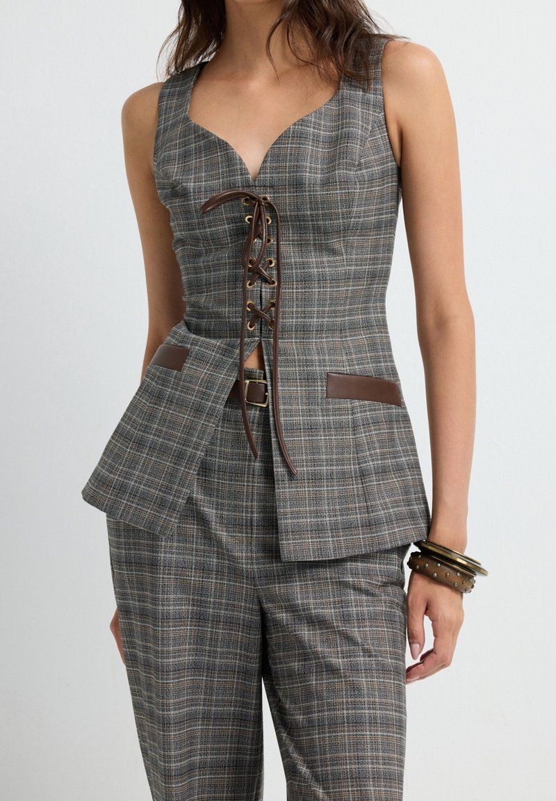 Femme portant un haut sans manches à carreaux gris avec un laçage marron à l'avant et un pantalon à carreaux assorti, accessoirisée avec des bracelets marron.