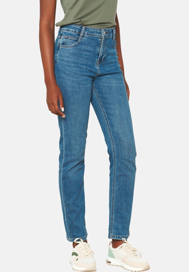 Šviesiai mėlyni džinsai su aukštu liemeniu, tiesiomis kojomis, šiek tiek blukimo ir penkių kišenių dizainu, derinami su žalia marškinėliais ir sportbačiais.