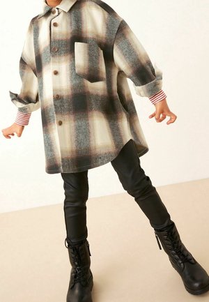 Lapsi, jolla on yllään liian iso beige-musta ruudullinen napillinen paita, mustat leggingsit, raidalliset hihansuut ja mustat nyörisaappaat, seisoo jalat levällään.