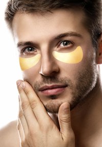 Onder-oogpatches van een zachte gele stof, geplaatst op de huid onder de ogen van een man die zijn gezicht aanraakt. Duidelijke huidstructuur.