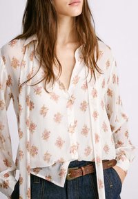 Camicia bianca trasparente con motivo floreale in rosa e oro, righe verticali, chiusura frontale con bottoni e vestibilità comoda. Abbinata a una cintura marrone.