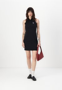 Vestido de polo sin mangas negro con cuello, que presenta un pequeño logo en el pecho. La modelo sostiene un bolso rojo y lleva zapatos negros y calcetines blancos.