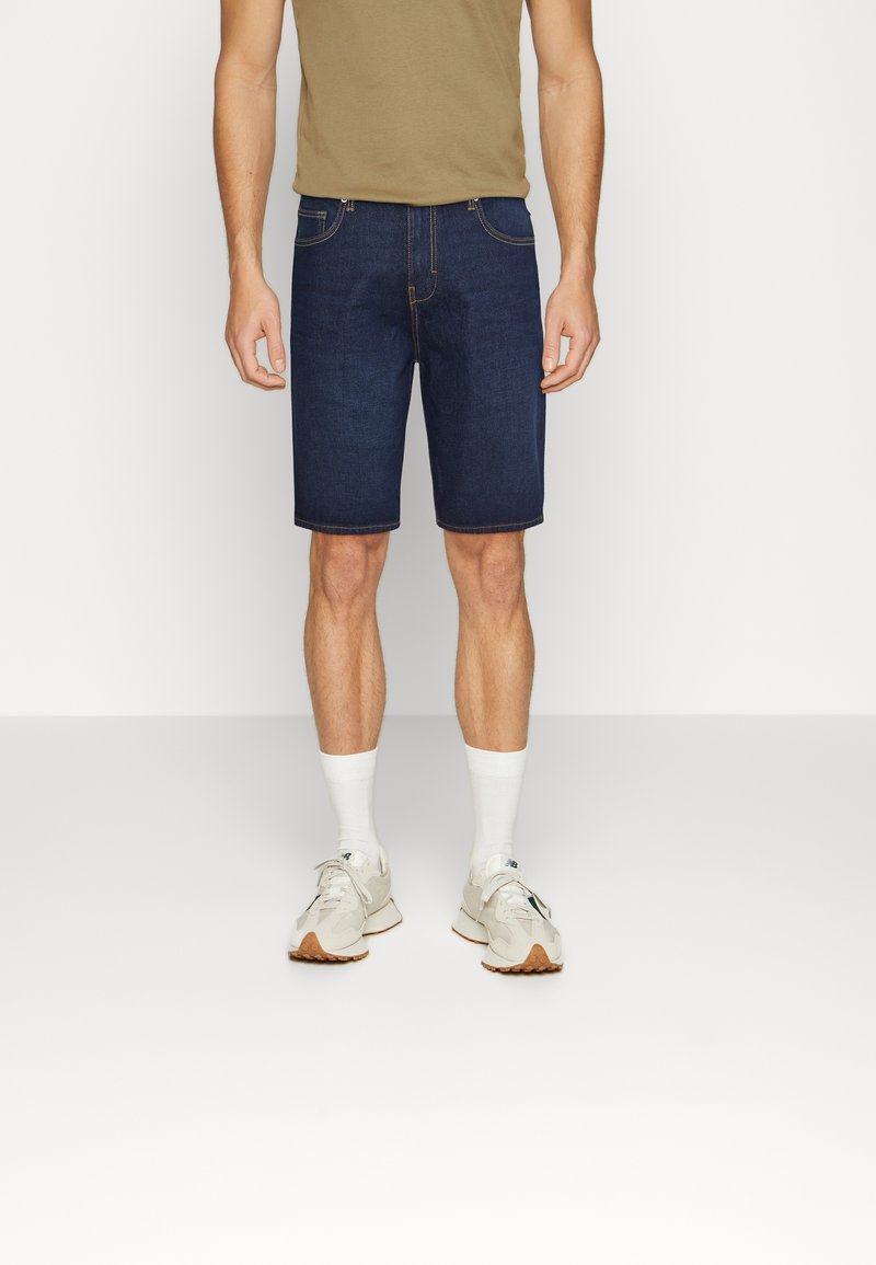 Homme portant un short en denim bleu foncé, un t-shirt beige, des chaussettes blanches et des baskets blanches, debout sur un fond blanc uni.