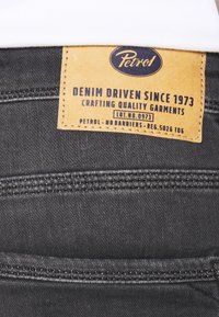 Tissu en denim noir avec une étiquette en cuir tan rectangulaire. L'étiquette présente un texte bleu indiquant "Petrol" ainsi que des instructions d'entretien. Coutures doubles visibles.