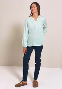 Mujer de pie junto a una pared beige, lleva una blusa de rayas verde claro, vaqueros azul oscuro y zapatos marrones, con una mano detrás de la espalda.