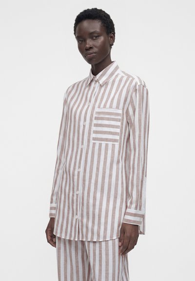 Γυναίκα που φοράει ένα μπεζ και λευκό πουκάμισο με κάθετες ρίγες και οριζόντια ριγέ τσέπη στο στήθος, συνδυασμένο με παντελόνι ίδιου σχεδίου.