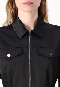 Camisa preta com fecho éclair, apresentando um colarinho pontudo, dois bolsos no peito com botões de pressão e detalhes de costura contrastantes. Textura de tecido suave.