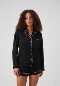 Svart pyjamas set bestående av en långärmad topp med vit piping och knappar, ihop med shorts som har kontrasterande fållar. Slät tygtextur.