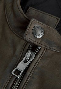 Brauner Lederjackenkragen mit einem runden Metallschnappverschluss, auf dem "SIGNATURE DURABLE GOODS" steht, und einem silbernen Reißverschluss mit einem rechteckigen Zieh-Tab.