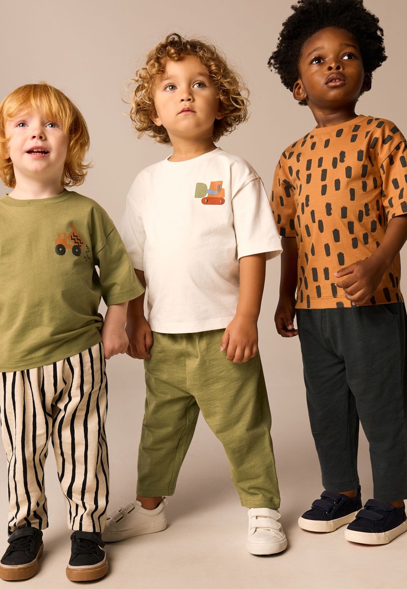 Drei Kinder tragen legere Outfits: ein grünes kurzärmeliges Hemd, ein weißes Hemd mit einem orangefarbenen Traktor-Motiv und ein braunes Hemd mit schwarzen Punkten; gestreifte und einfarbige Hosen.