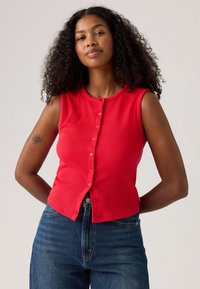 Haut sans manches rouge à côtes avec un col rond et des détails de boutons à l'avant. Associé à un jean en denim bleu foncé. Tatouage visible sur le bras.