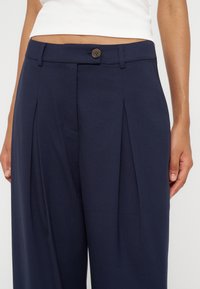 Personne portant un pantalon plissé bleu marine taille haute avec un bouton et des passants de ceinture, associé à un top court blanc.