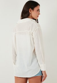 White long-sleeve shirt made of lightweight, slightly textured fabric. Features a classic collar, buttoned cuffs, and a curved hem.