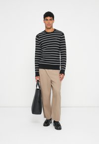 Maglione a righe bianche e nere, con polsini e orlo a costine, pantaloni beige, in mano una borsa nera, con texture liscia e vestibilità rilassata.