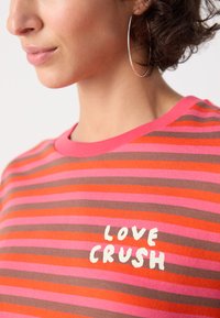 Raidoitu t-paita vaaleanpunaisena, punaisena ja ruskeana, jossa on pyöreä kaula-aukko. Keskellä on valkoiset tekstit "LOVE CRUSH". Pehmeää puuvillakangasta.